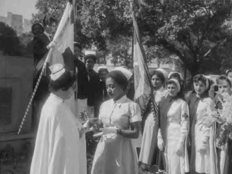 Uma professora, provavelmente, entrega algo não identificado a uma aluna negra. Ao fundo, diversas outras alunas, todas com uniforme de enfermeira. Uma segura a bandeira do Brasil. Outra segura uma bandeira não identificada.