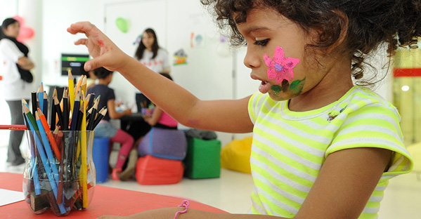 Criança brincando com lápis de cor. Em seu rosto, está pintada uma flor.