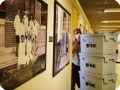 Homem conduz pilha de caixas. Ele adentra um corredor em cujas paredes estão fotos históricas em preto e branco.