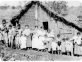 Família composta de homens, mulheres e crianças, reúne-se em frente a uma casa de pau-a-pique coberta com sapê